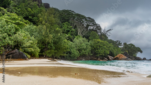 The landscape of a tropical island. The green hill is overgrown with lush vegetation. A pile of boulders by the water. The waves of the turquoise ocean are foaming on the beach. Wet sand glistens.