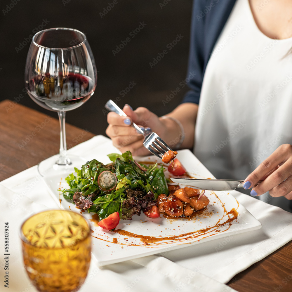 Woman is having salad and glass of red wine for lunch. Knife and fork in hands of woman. Lunch at table in restaurant. Vegetable salad on white plate onawooden table. Soft focus. 