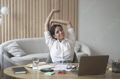 Tired italian lady remote employee stretching arms while sitting at desk with laptop