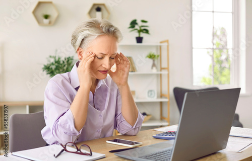 Unhappy gray-haired adult woman suffers from headaches and migraines and hold on head due to regular overwork and excessive stress sits at table with laptop in office. Professional burnout concept