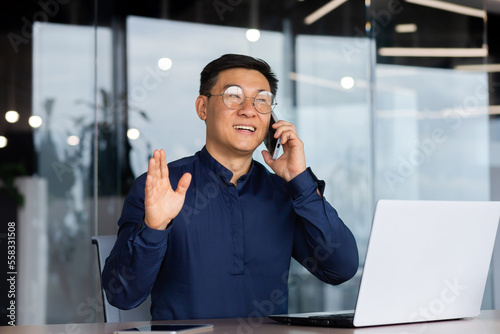 Successful and cheerful asian businessman talking on the phone with clients inside the office, mature man in a shirt working on a laptop at the workplace.