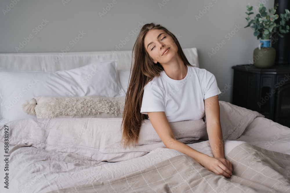 Sleepy blonde caucasian young woman in white t-shirt sitting on bed eyes closed awakening cute smiling at Sunday morning. Relaxed Italian girl with long loose hair enjoys weekend at cozy home. Sleepy blonde caucasian young woman in white t-shirt sitting on bed eyes closed awakening cute smiling at Sunday morning. Relaxed Italian girl with long loose hair enjoys weekend at cozy home.
