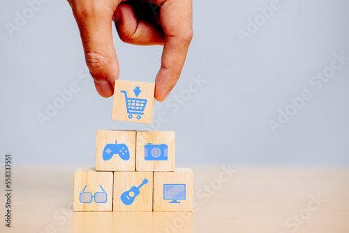Hand holding wooden cube with cart icon joystick, camera, glasses, guitar, computer. The concept of shopping, buying items and goods.