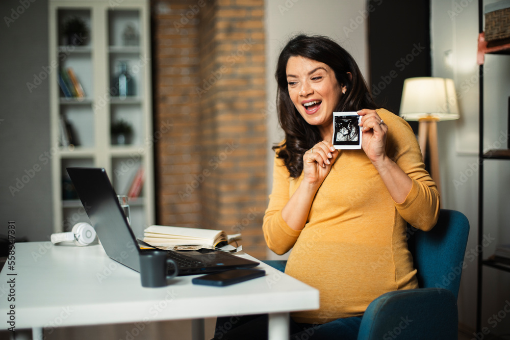 Beautiful young pregnant woman looking at ultrasound pictures of her baby. Businesswoman in office Beautiful young pregnant woman looking at ultrasound pictures of her baby. Businesswoman in office