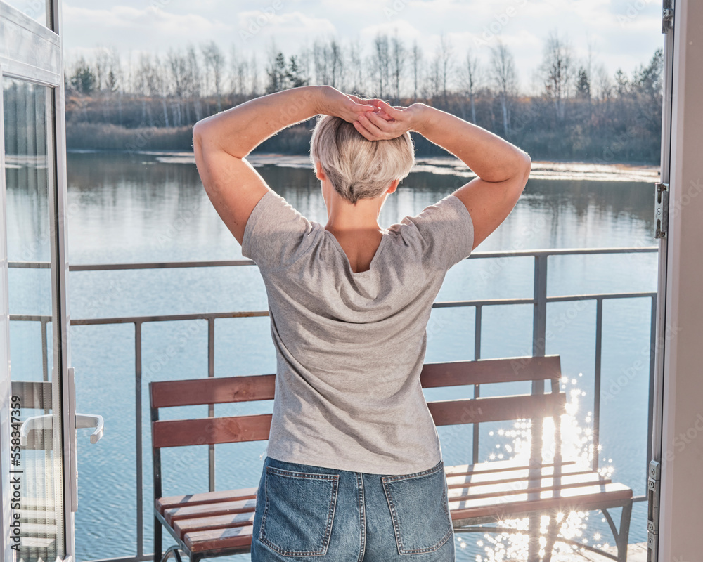 Senior woman with short gray hair stretching oneself on veranda of country house on lake shore.