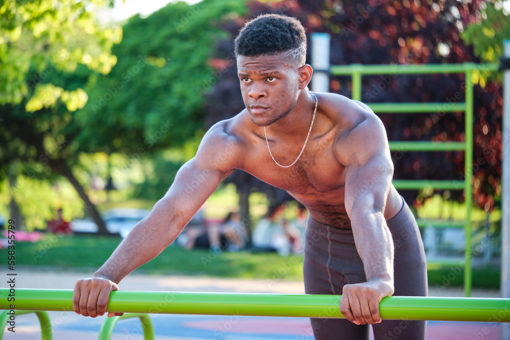 Obraz premium Young fit shirtless black man doing push-ups on a bar in a calisthenics park outdoors on sunny day. Fitness and sport lifestyle.