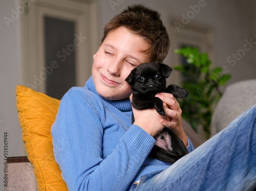 Boy playing with baby dog. Kid play with puppy at home. Little boy and griffon or brabanson dog on sofa. Children and pets at home. Kid sitting on the floor with pet. Animal care. photo