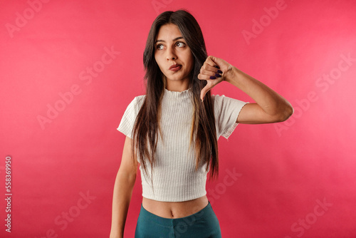 Young caucasian woman wearing casual top isolated over red background showing a thumbs down expression with a puzzled look. Not satisfied and looks at the camera.