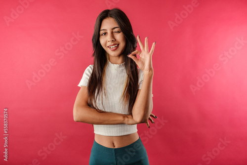 Young beautiful woman wearing ribbed crop isolated over red background totally agree with you  showing okay sign and nod approval. Blinking and everything s fine.