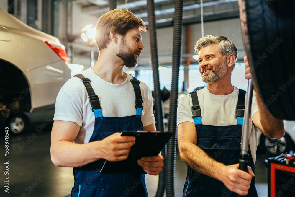 Obraz premium Automotive breakdown. Cheerful experts at work shop, standing in blue safety uniform, white t shirt, hold pda device, background of vehicle.