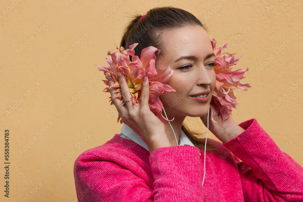 Fototapeta premium Smiling brunette woman in pink sweater wearing floral headphones and listening music. Dahlia flower buds as a earphones.