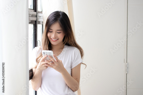 Portrait of attractive asian woman in casual clothes is using a smart phone and smiling while standing near the window, holding mobile phone.