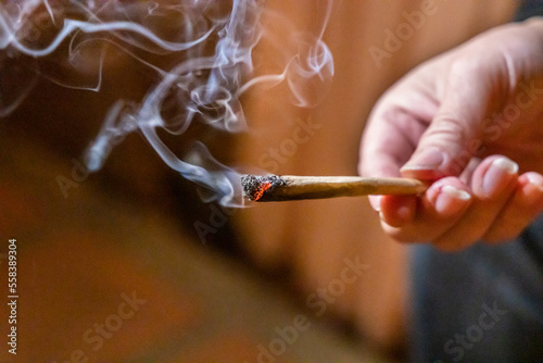 Finger holding lighted cannabis cigarette with smoke in dark room, with selective focus on cannibis ash and fire.