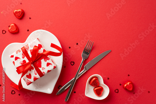 Valentine's Day concept. Top view photo of heart shaped plates with large giftbox knife fork chocolate candies and confetti on isolated red background with blank space