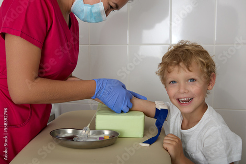 Cheerful boy bravely gave his hand to a nurse in a red robe to draw blood. Cute child smiles while a laboratory employee performs manipulations to take blood from a vein photo