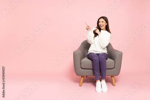 Young elegant Asian woman smiling and pointing to empty copy space and sitting on armchair isolated on pink background
