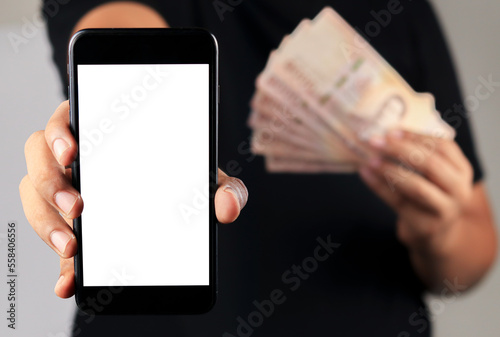 Fototapeta Asian man stand and holding smart phone screen another hand showing Thai baht money over grey background