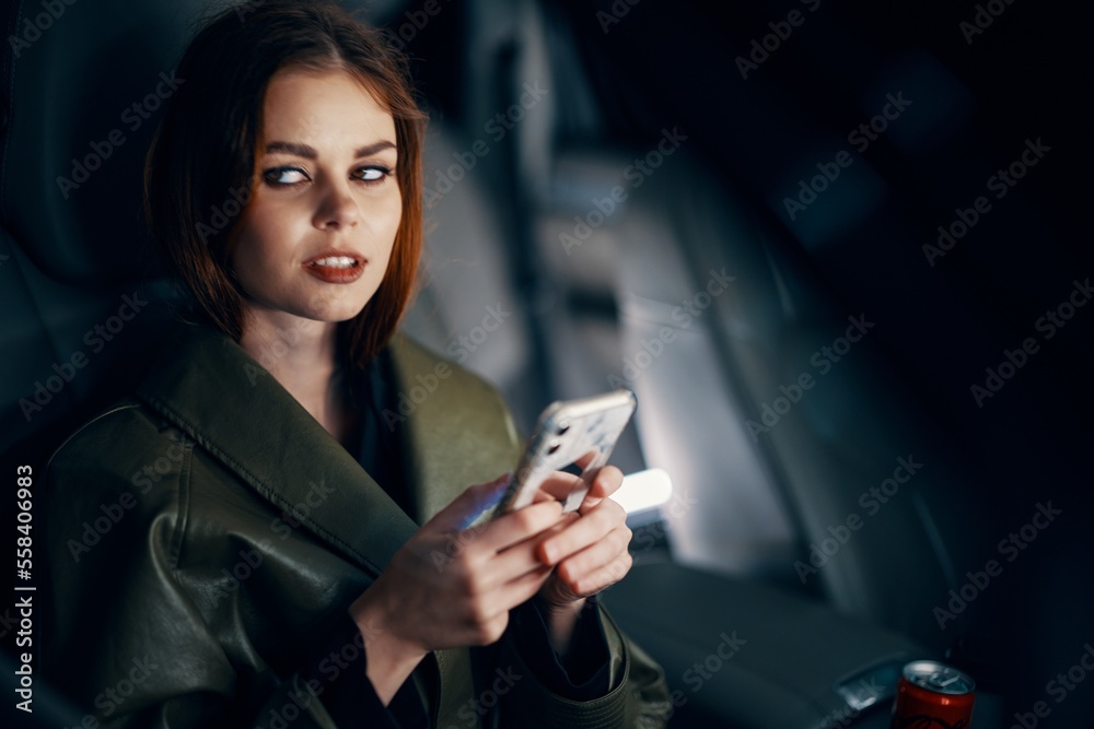 a close horizontal portrait of a stylish, luxurious woman in a leather coat sitting in a black car at night in the passenger seat, looking pleasantly into the camera holding a smartphone in her hand