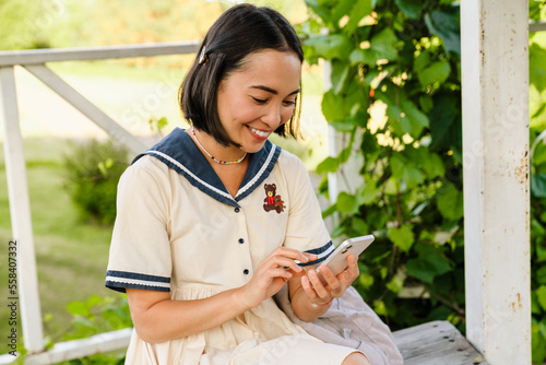 Young cheerful asian girl using mobile phone while sitting on bench in park