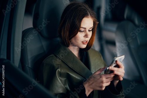 a close horizontal portrait of a stylish, luxurious woman in a leather coat sitting in a black car at night in the passenger seat, looking at her phone during the trip © SHOTPRIME STUDIO