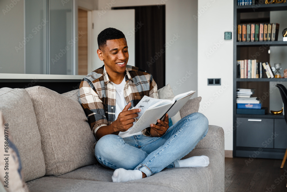 Obraz premium Young smiling african man reading book while sitting on couch at home