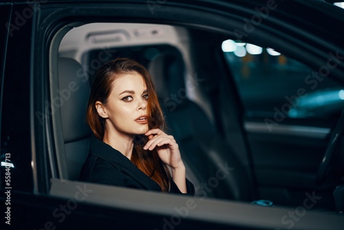 horizontal photo from the side, at night, of a woman sitting in a black car and thoughtfully looking at the camera holding her hand near her face, parking the car in the parking lot