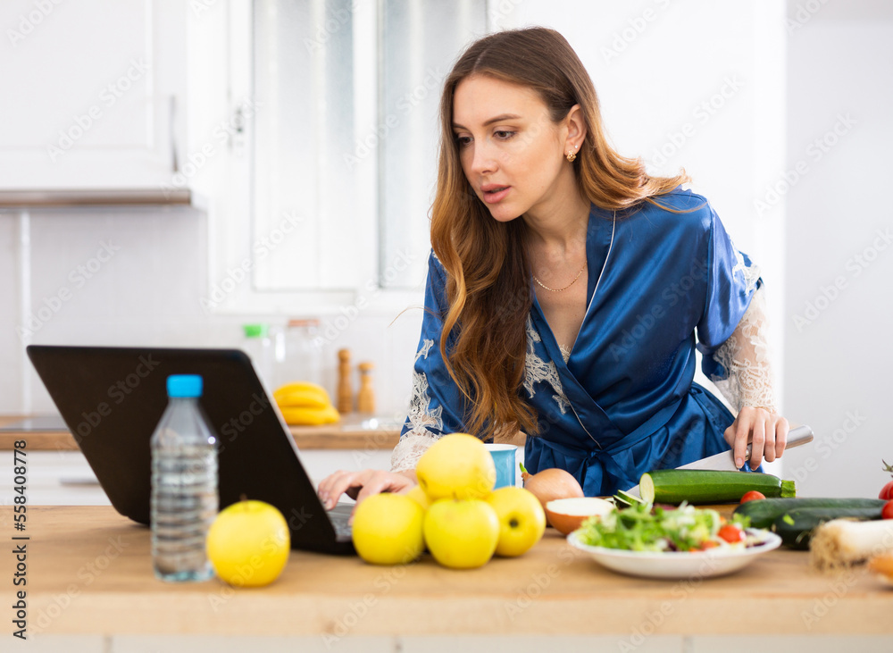 Housewife in housecoat reads recipe in laptop and prepares a salad in kitchen