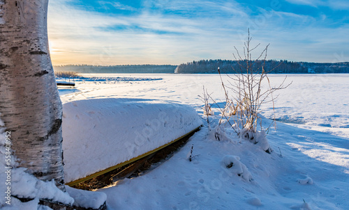 Frozen lake with lot of snow and rowboat upside down on shore with snow on top and a birch tree