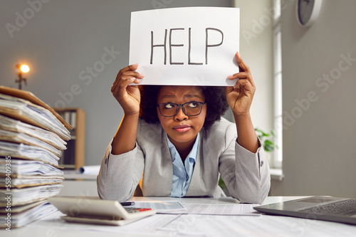 Tired, busy, sad African American woman in a suit and glasses sitting at an office desk with a load of paperwork and holding a paper sign with the word HELP Fototapeta