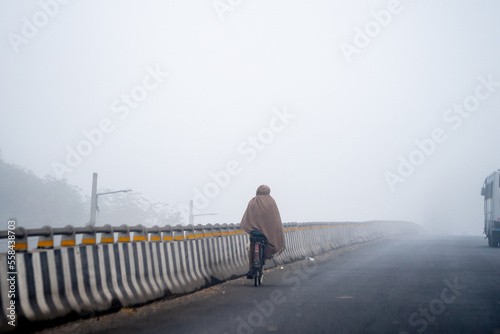 Indian man riding on bicycle in the cold winter morning with dense fog wrapped up tightly showing the harsh winters of Delhi, Rajasthan and Haryana photo