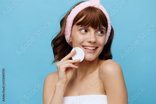 a cute woman with even radiant skin and dark silky beautiful hair stands on a blue background in a white towel with a pink bandage on her head after a bath, removes makeup from her face. Horizontal