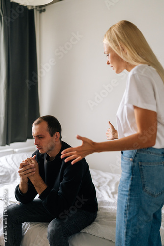 Vertical portrait of stressed depressed man sitting on bed looking away ignoring angry blonde wife arguing blaming upset man of problems at home. Jealous girlfriend shouting at sad boyfriend.
