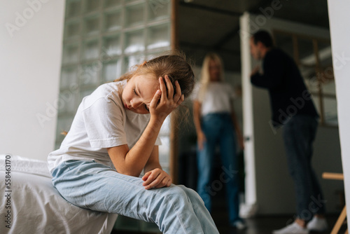 Front view of sad lonely little girl looking away, crying suffering sitting on sofa during parents quarrelling and fighting in living room on background. Concept of family problems, conflict, crisis.