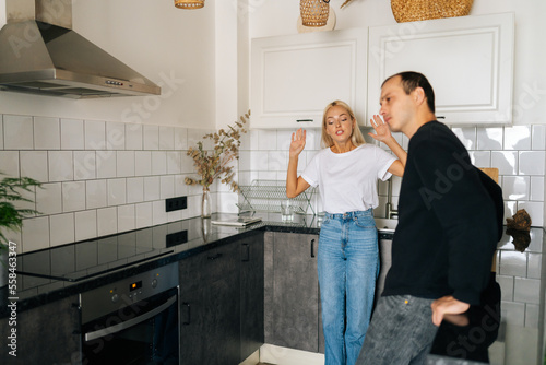 Jealous girlfriend scolding, raising voice, gesturing with hands, yelling, having argument with ignoring tired boyfriend standing in kitchen. Concept of family crisis, domestic violence, abuse