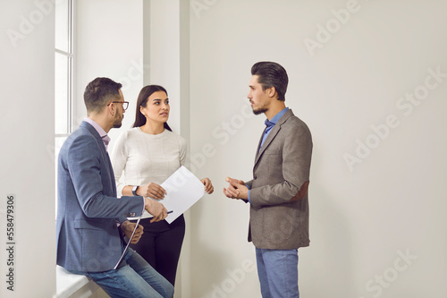 Group of office workers having a discussion. Team of three people talking about something while standing together by an office window. Work, business concepts