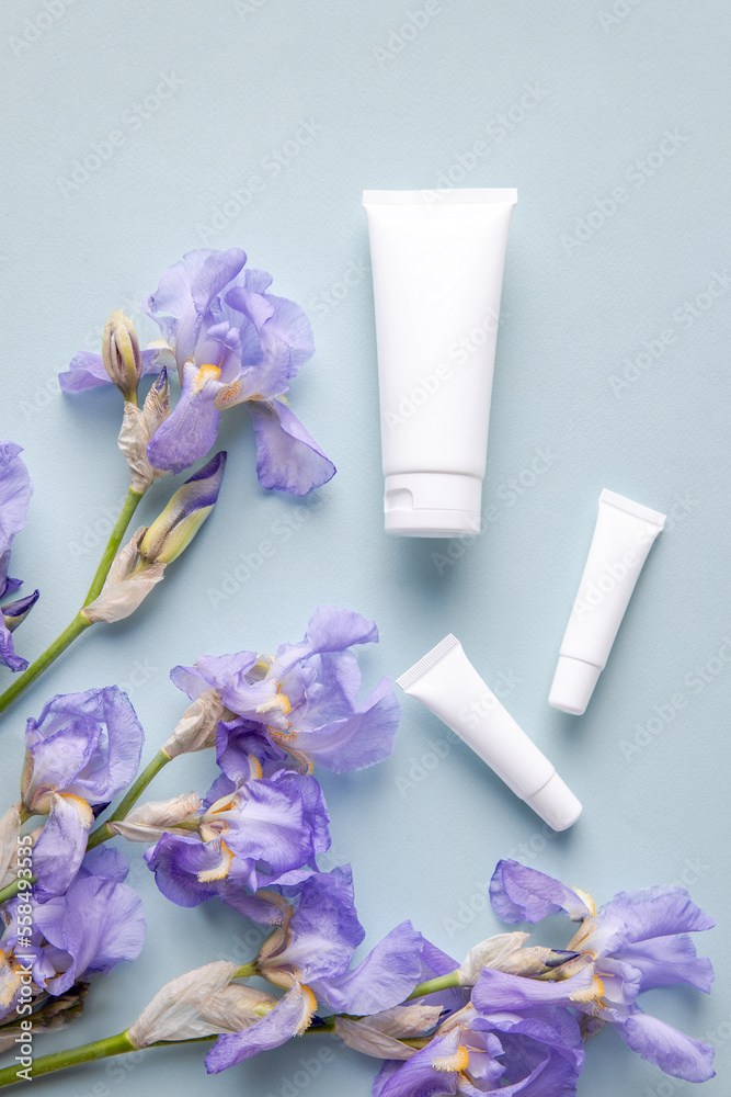Mockup for a cosmetic brand. A set of White jars of SPA cosmetics on a blue background, surrounded by natural purple iris flowers. The concept of eco products, minimalist style. Place for text Mockup for a cosmetic brand. A set of White jars of SPA cosmetics on a blue background, surrounded by natural purple iris flowers. The concept of eco products, minimalist style. Place for text