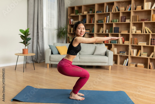 Happy fit japanese lady exercising in living room alone, doing squats, working on buttocks, smiling at camera