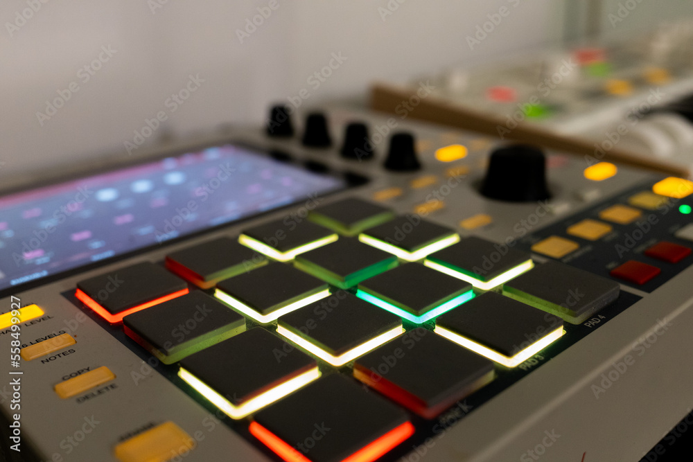 Drum machine with colored lights on pads and display. Piano keyboard and knobs blurred for focus on pads. 