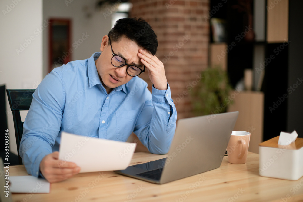 Confused asian mature man sitting in front of laptop, reading papers and touching his head, copy space Confused asian mature man sitting in front of laptop, reading papers and touching his head, copy space