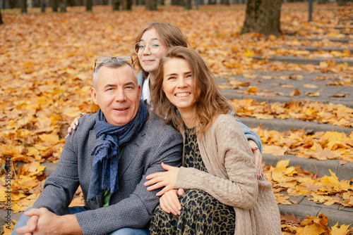 Portrait of family embracing  sitting on concrete stairs covered with yellow fallen leaves among trees in park forest.
