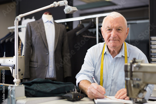 Elderly man tailor takes measurements from his jacket and writes them down in notebook in a workshop