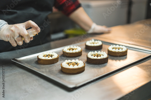 A expert wearing transparent gloves, black apron and red plaid shirt spraying tarts decorated with white cream and golden pearls on a baking tray. Blurred background. High quality photo