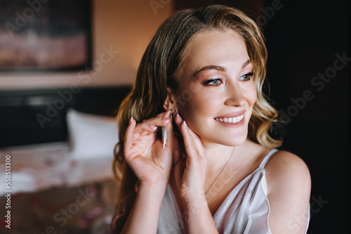 A beautiful curly blonde bride in a white dress straightens an earring on her ear while standing in the room. Wedding photography  close-up portrait.