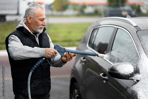 Side portrait of elderly man driver, spraying jet of water on car from high pressure vehicle washer