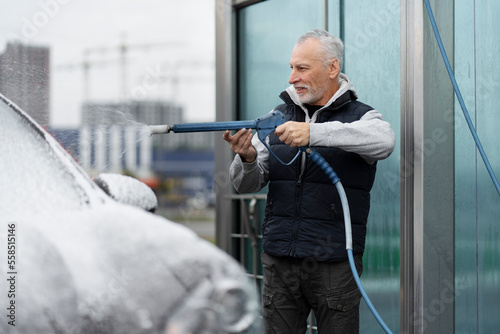 Caucasian bearded grey-haired elderly man, a driver using a pressurized water hose sprays a jet of water to clean the car from cleaning foam while washing his automobile, at a self-service car wash