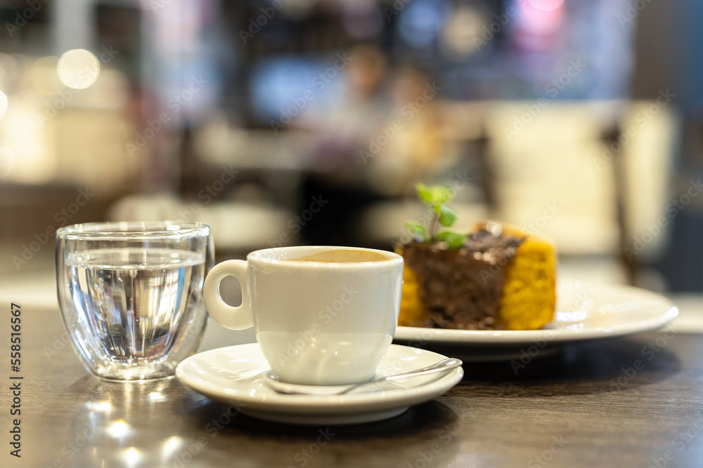 A mug of coffee, a glass of water and a chocolate cake with mint leaves on a wooden table in a Brazilian cafe. Blurred people in the background