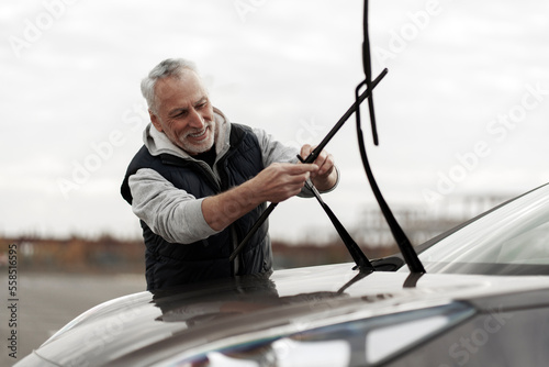 Handsome smiling senior Caucasian man, driver installing new windshield wipers by himself at outdoors parking lot. Automobile industry. Safety driving. Car maintenance. People. Auto service concept 
