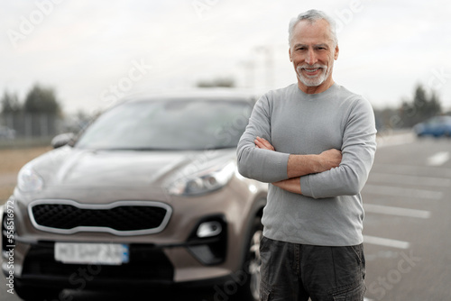 Handsome elderly man driver, smiles at camera, stands with arms crossed near his automobile outdoors
