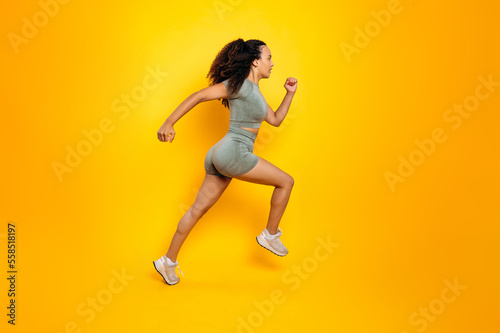 Full length side view photo of a fitness sporty brazilian or hispanic woman in sportswear, training working out, jumping, running, looking aside, isolated orange background. Sports lifestyle