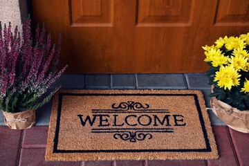 Door mat with word Welcome and beautiful flowers near entrance